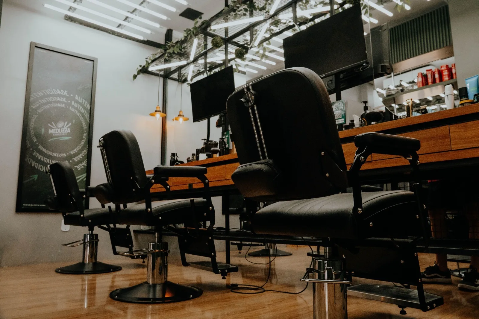 Salão da Meduzza Barbearia: três cadeiras de barbeiro pretas alinhadas sobre piso de madeira, bancada de madeira com produtos e luminárias ao fundo.