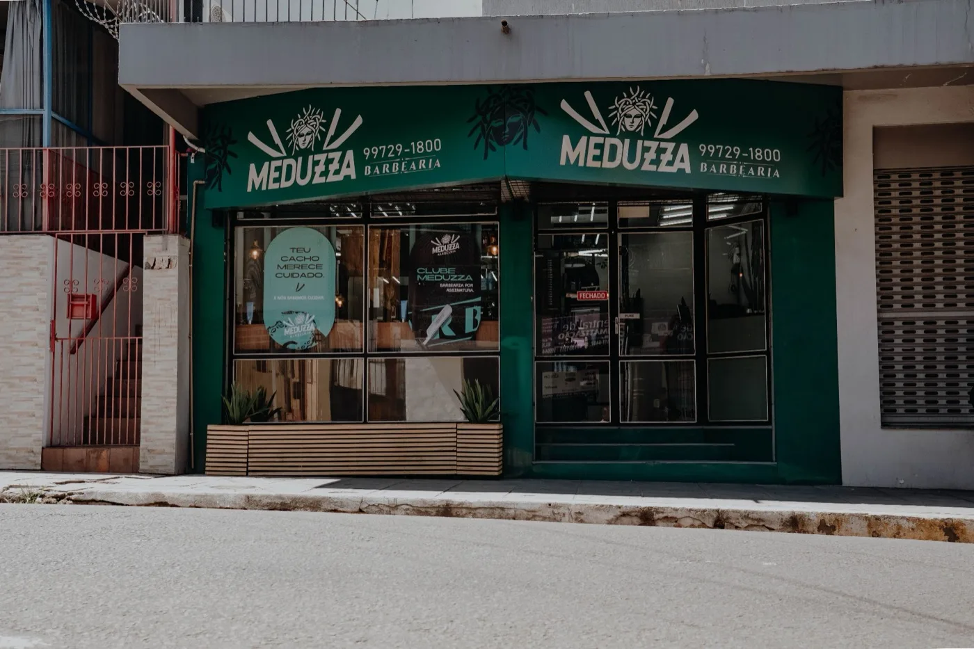 Fachada da Meduzza Barbearia: toldo verde-escuro com a marca Meduzza e o telefone 99729-1800, vitrine de vidro com adesivos e bancos de madeira na calçada.